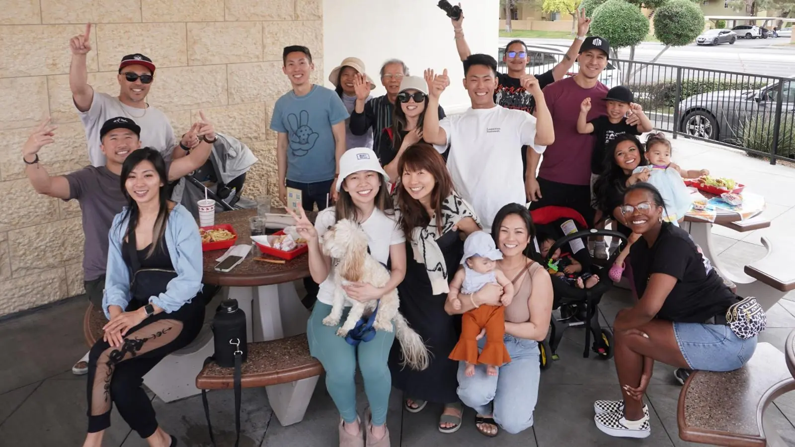 A Friends Club social: friends, families, babies and a dog gathered around patio tables, waving and smiling