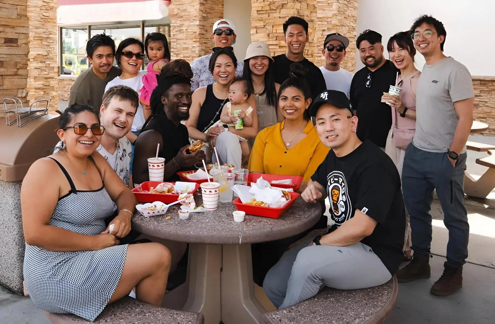 Friends gathered around a patio table, sharing food and laughing, a baby on one lap